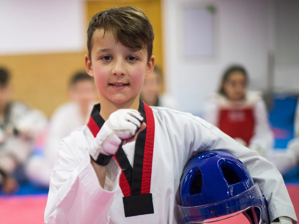 A young student in a dobok holding a sparring helmet at the side of the mat